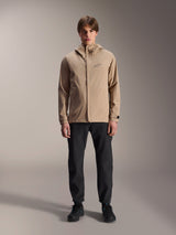 Alpinestars A-Dura Rain Jacket, Light Brown cycling jacket, worn with black pants and black shoes, featuring a small chest logo and hood, professional mountain bike apparel for all-weather riding.