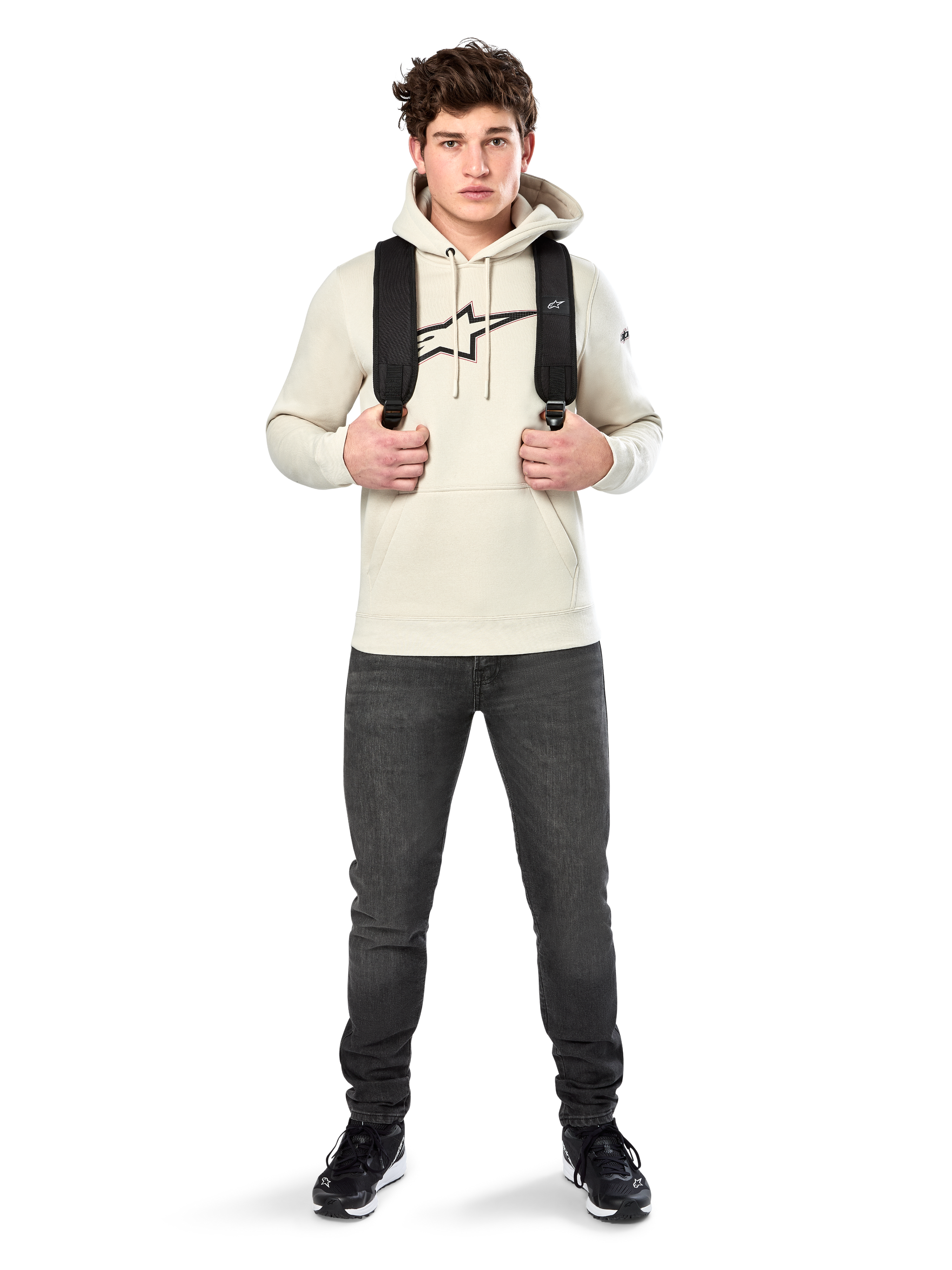 Alpinestars Insignia Hoodie, casual streetwear, Off White fleece pullover, carbon fiber look logo with red outline, drawstring hood, kangaroo pocket, worn by model with black backpack and dark grey jeans