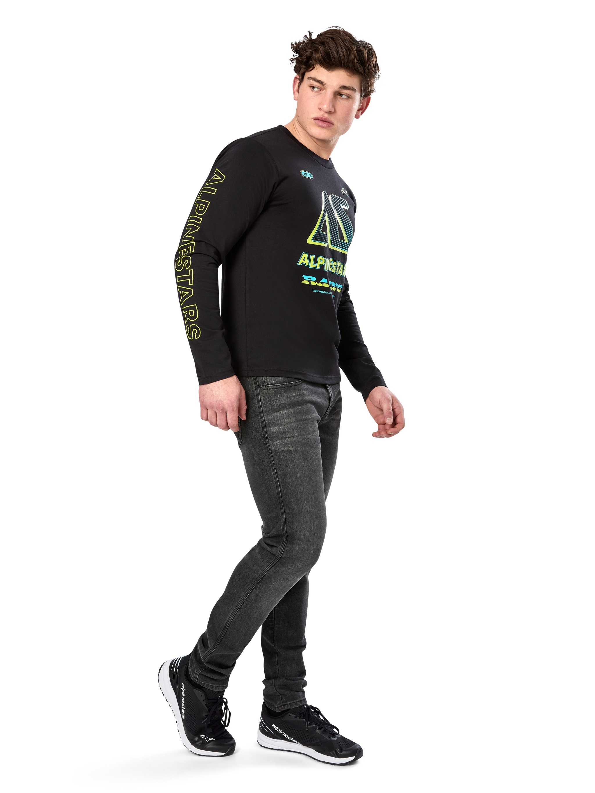 Man wearing Alpinestars Ayess CSF Long Sleeve Tee, Black, featuring yellow sleeve lettering and vibrant lime and cyan chest graphics, paired with dark grey jeans and black sneakers, casual motorsport-inspired streetwear.
