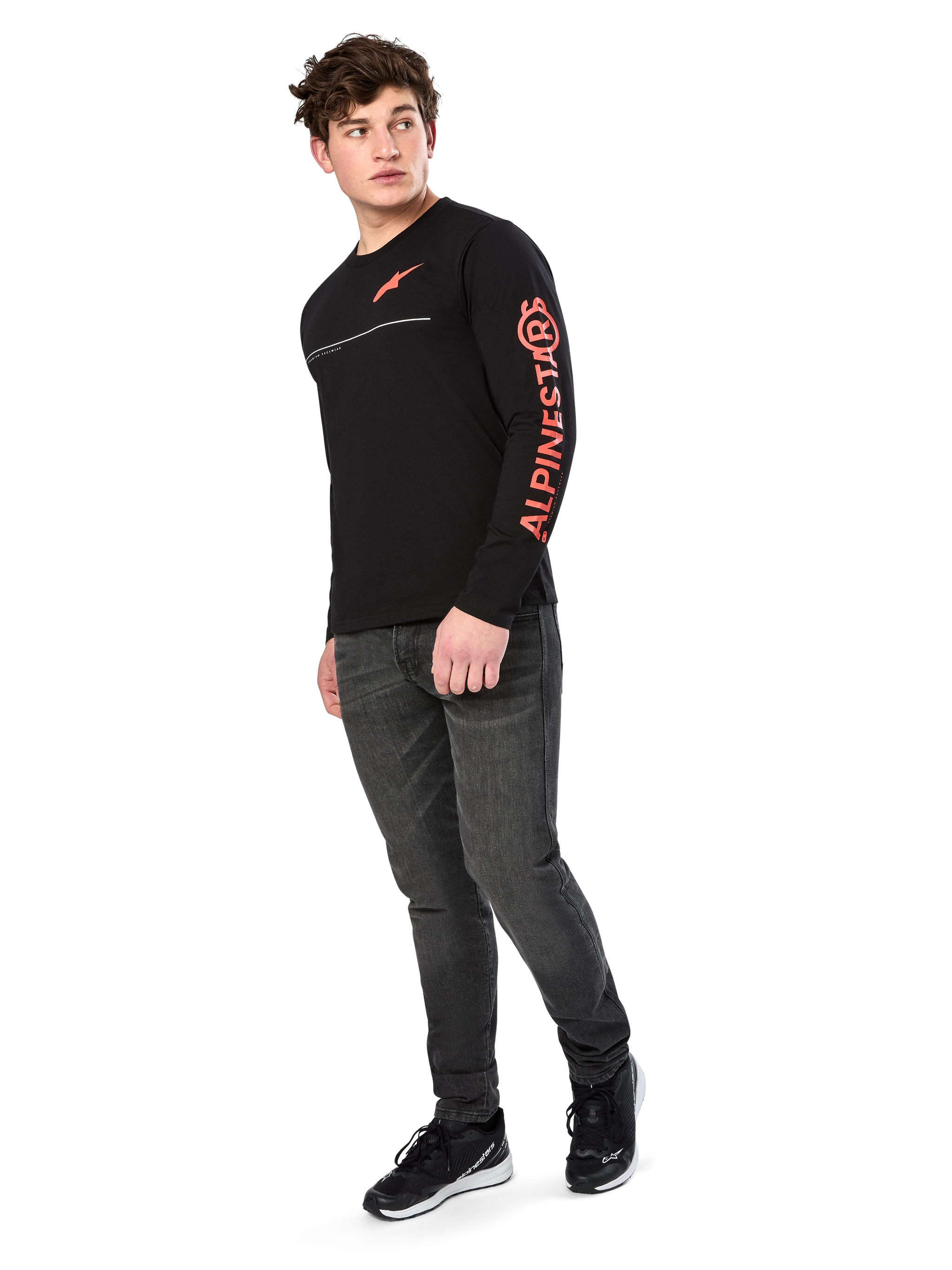 Male model wearing Alpinestars Committed CSF Tee, Long Sleeve T-Shirt, Black, orange-red chest logo and sleeve branding, paired with dark gray jeans and black sneakers, casual motorsport-inspired streetwear