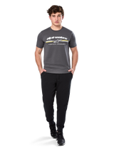 Man wearing Alpinestars Prevail Csf Tee, Short Sleeve T-Shirt, Charcoal, featuring white Alpinestars logo and tri-color stripe with yellow accents, paired with black joggers and sneakers, casual motorsport streetwear