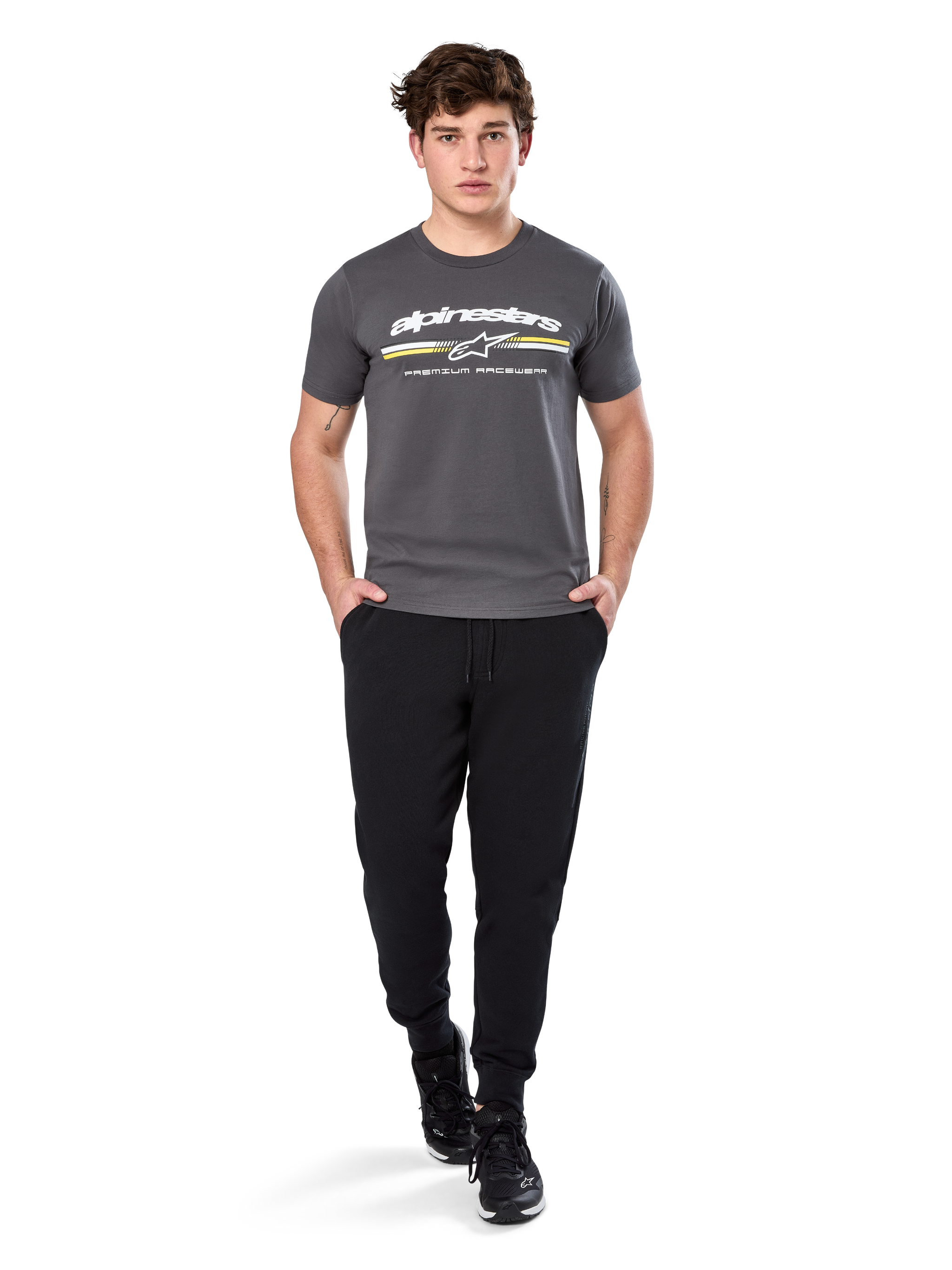 Man wearing Alpinestars Prevail Csf Tee, Short Sleeve T-Shirt, Charcoal, featuring white Alpinestars logo and tri-color stripe with yellow accents, paired with black joggers and sneakers, casual motorsport streetwear