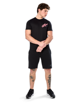 Model wearing Alpinestars Attrition Performance Tee, Short Sleeve Performance T-Shirt, Black with a pink Astars logo on the chest, paired with black shorts and sneakers, casual motorsport-inspired sportswear