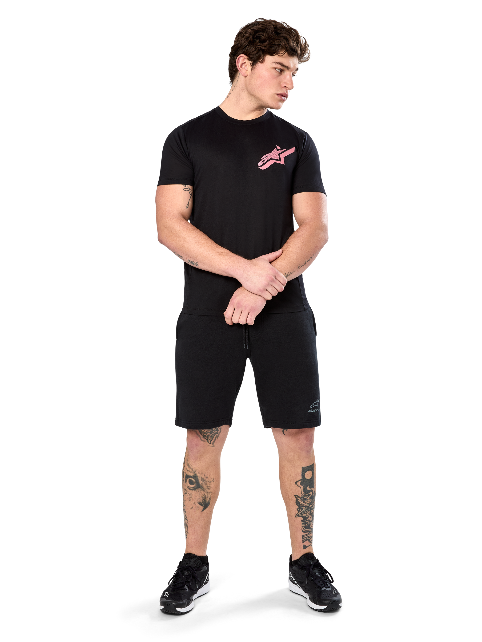Model wearing Alpinestars Attrition Performance Tee, Short Sleeve Performance T-Shirt, Black with a pink Astars logo on the chest, paired with black shorts and sneakers, casual motorsport-inspired sportswear