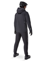 Alpinestars A-Dura Elite Waterproof Jacket, Black, rear view showing over-the-helmet hood and longer drop tail hem, paired with matching black MTB pants, gloves, and helmet for complete all-weather trail protection.