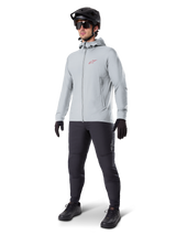 Alpinestars A-Dura Thermal Jacket in Titanium, light gray hooded cycling jacket with a red Alpinestars logo on the chest, worn by a man with a black MTB helmet, sunglasses, dark gray pants, and black gloves, professional mountain bike gear