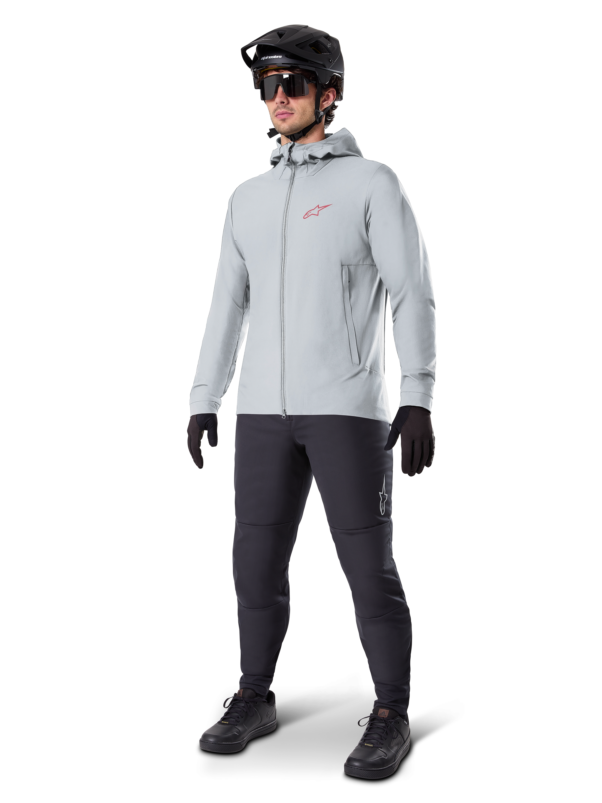Alpinestars A-Dura Thermal Jacket in Titanium, light gray hooded cycling jacket with a red Alpinestars logo on the chest, worn by a man with a black MTB helmet, sunglasses, dark gray pants, and black gloves, professional mountain bike gear