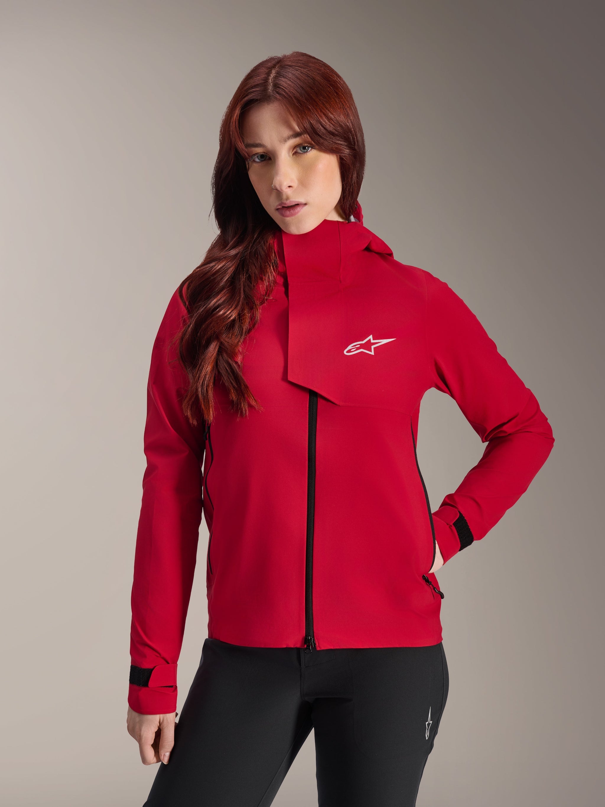 Alpinestars Stella A-Dura Elite Waterproof Jacket, Women's Mountain Bike Jacket, Red Fluo, featuring a front storm flap with a white Alpinestars logo, high collar, and adjustable cuffs, worn with black pants for a technical riding look