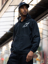 Alpinestars Speeder Chromium Windbreak Jacket, Black, hooded windbreaker with a white chest logo, worn by a man with a matching black Alpinestars cap in an urban setting, casual motorsport streetwear.