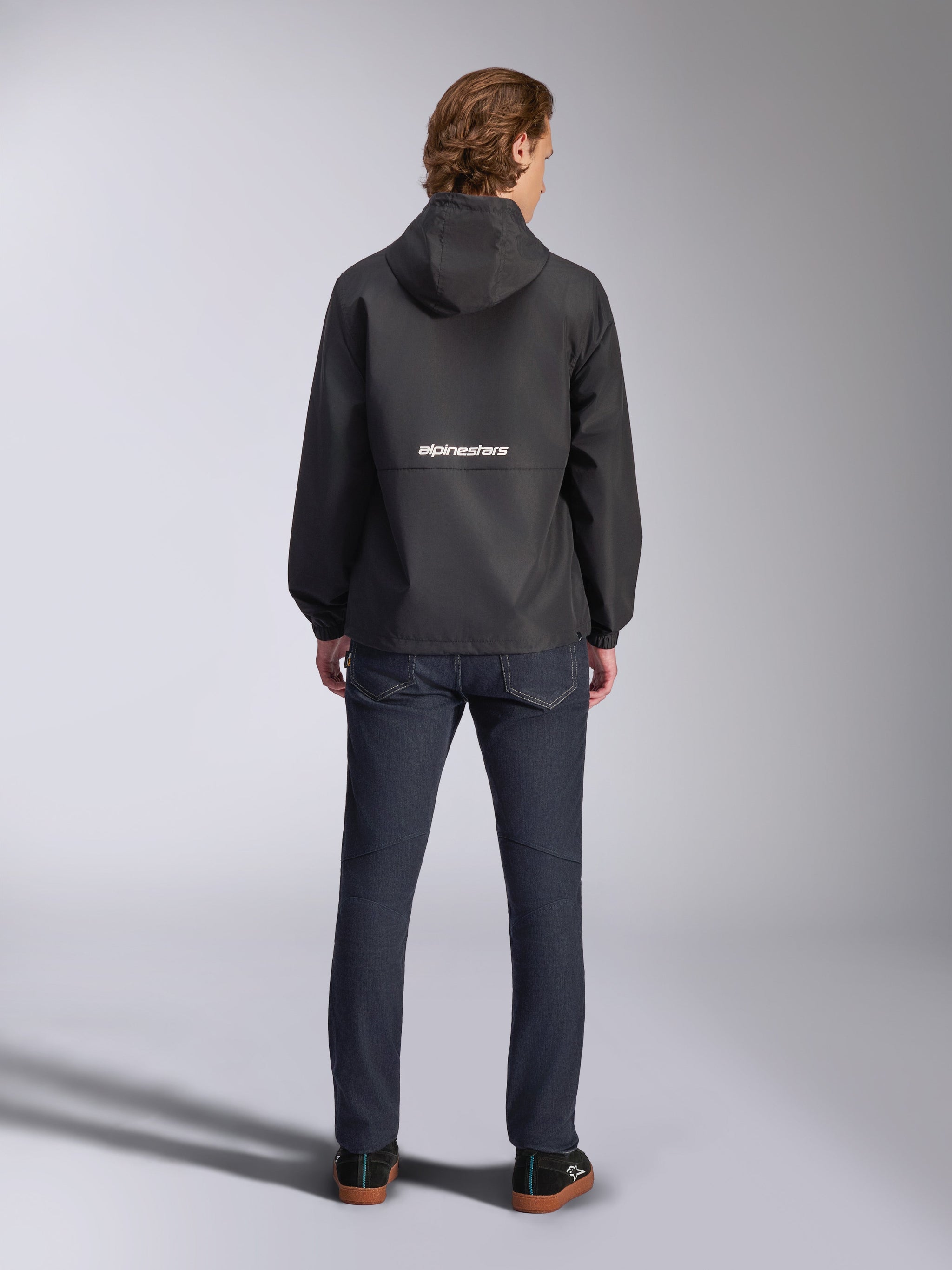 Alpinestars Speeder Chromium Windbreak Jacket, Black, back view with white Alpinestars branding across the middle, hooded style, worn by model with dark denim pants, casual motorsport-inspired jacket