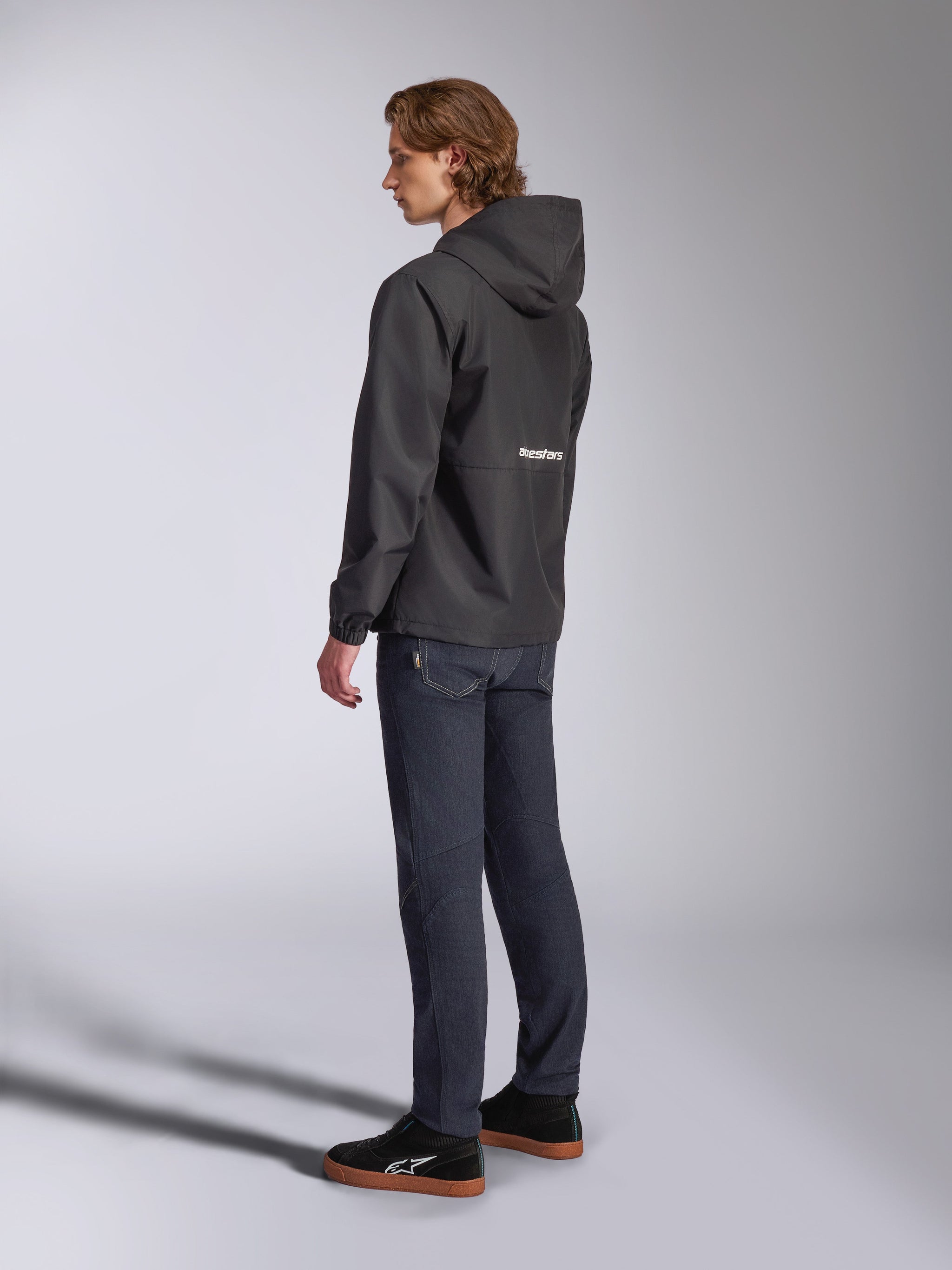 Alpinestars Speeder Chromium Windbreak Jacket, Black with white logo on the back, men's casual windbreaker with hood, side profile view worn with dark jeans and black sneakers, motorsport-inspired streetwear