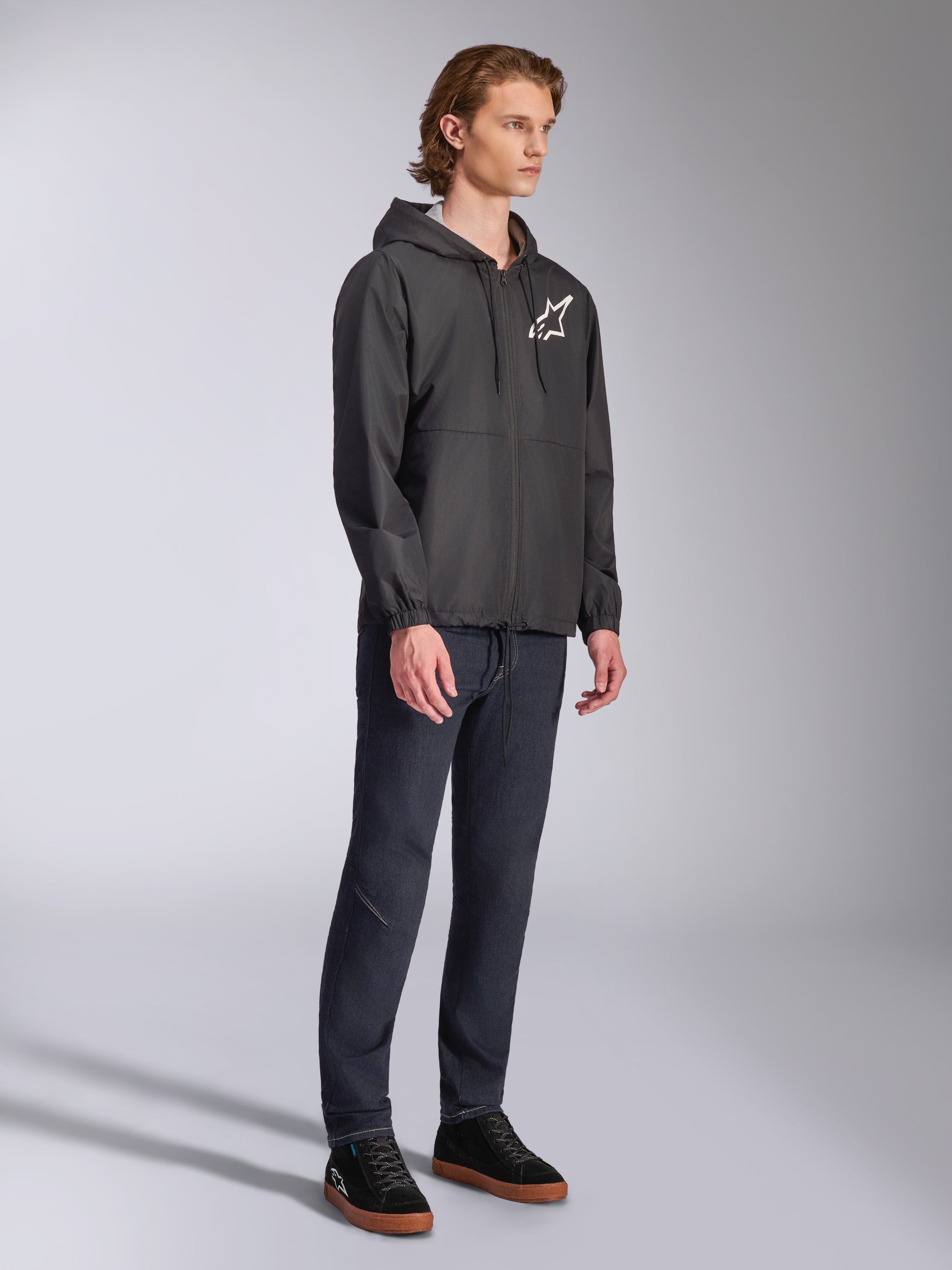 Alpinestars Speeder Chromium Windbreak Jacket, Black, hooded windbreaker with front zipper, white Alpinestars star logo on the left chest, casual motorsport streetwear, styled with dark pants and black sneakers in a studio setting.