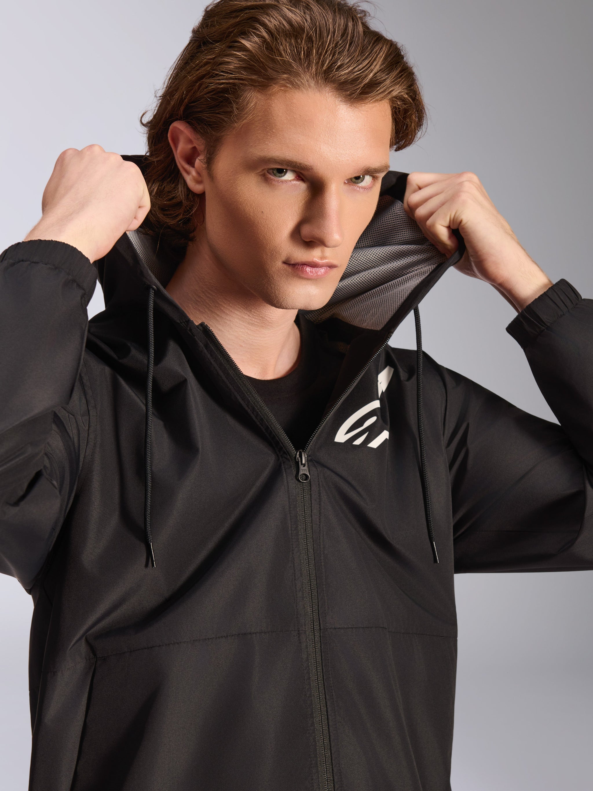 Alpinestars Speeder Chromium Windbreak Jacket, Black, worn by a male model holding the hood, features a white logo on the chest, mesh hood lining, and full-zip front, casual motorsport-inspired streetwear