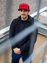 Man wearing the Alpinestars Overhaul Racer Jacket in Black, a hooded water-resistant rain shell jacket with a small chest logo, accessorized with a red Alpinestars cap, casual motorsport lifestyle apparel