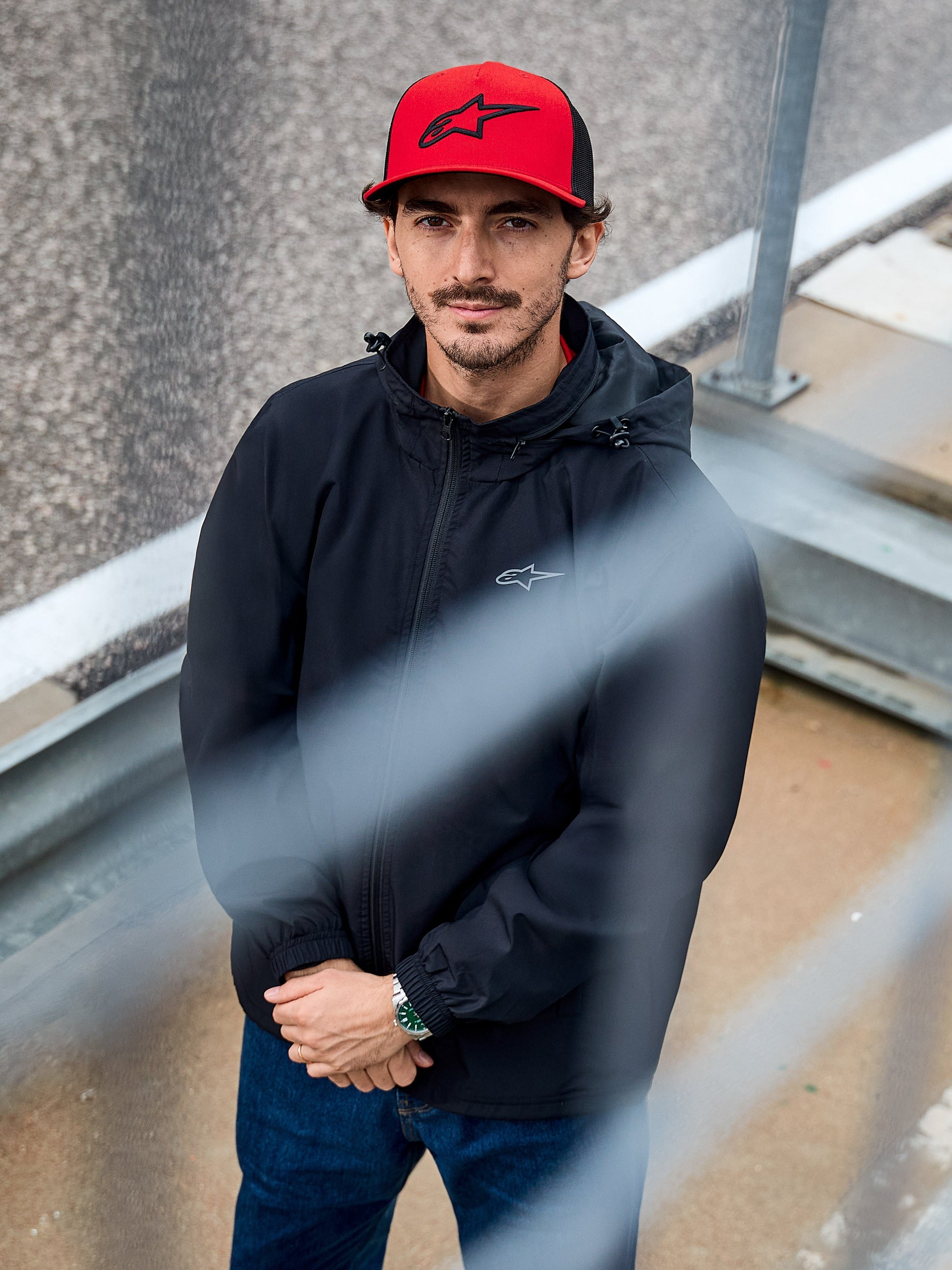 Man wearing the Alpinestars Overhaul Racer Jacket in Black, a hooded water-resistant rain shell jacket with a small chest logo, accessorized with a red Alpinestars cap, casual motorsport lifestyle apparel