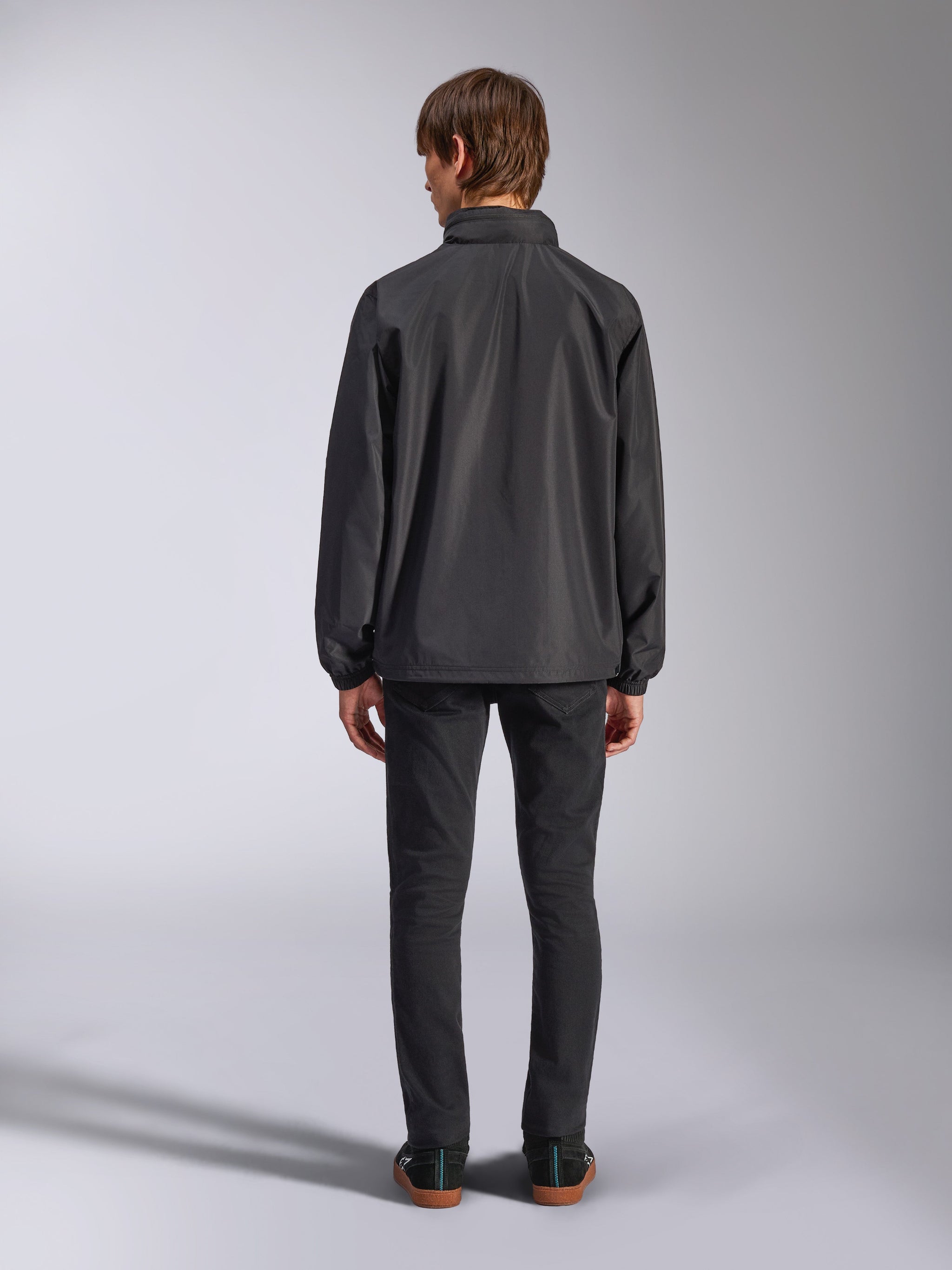 Alpinestars Overhaul Racer Jacket, Black, back view of a man wearing the rain shell jacket, features water-resistant 2.5L fabric, elasticated cuffs, and fixed hood, paired with black pants, casual motorsport-inspired outerwear