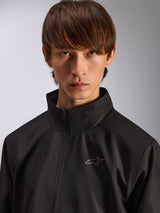 Alpinestars Overhaul Racer Jacket, Black rain shell jacket, close-up portrait of a male model wearing the jacket, featuring a high collar and small silver Astars logo on the chest, motorsport-inspired casual outerwear