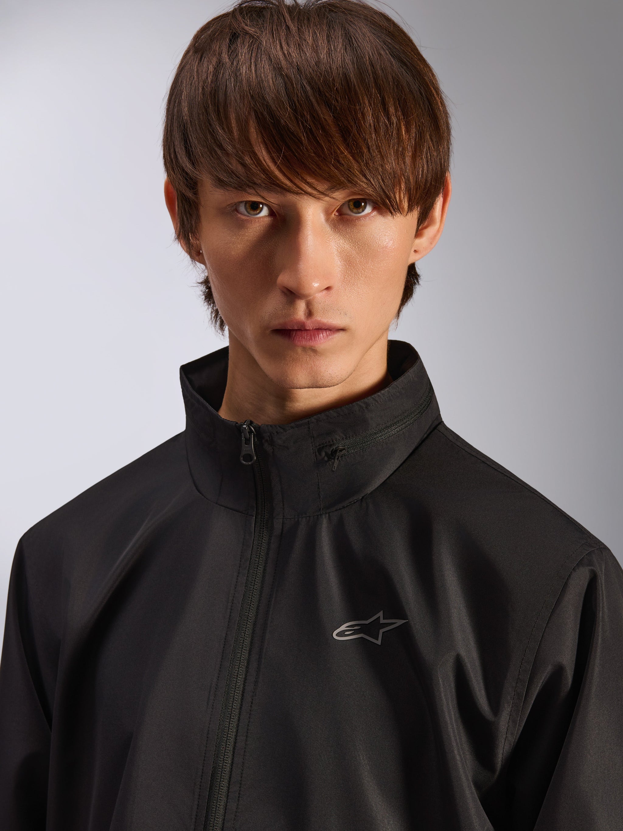 Alpinestars Overhaul Racer Jacket, Black rain shell jacket, close-up portrait of a male model wearing the jacket, featuring a high collar and small silver Astars logo on the chest, motorsport-inspired casual outerwear