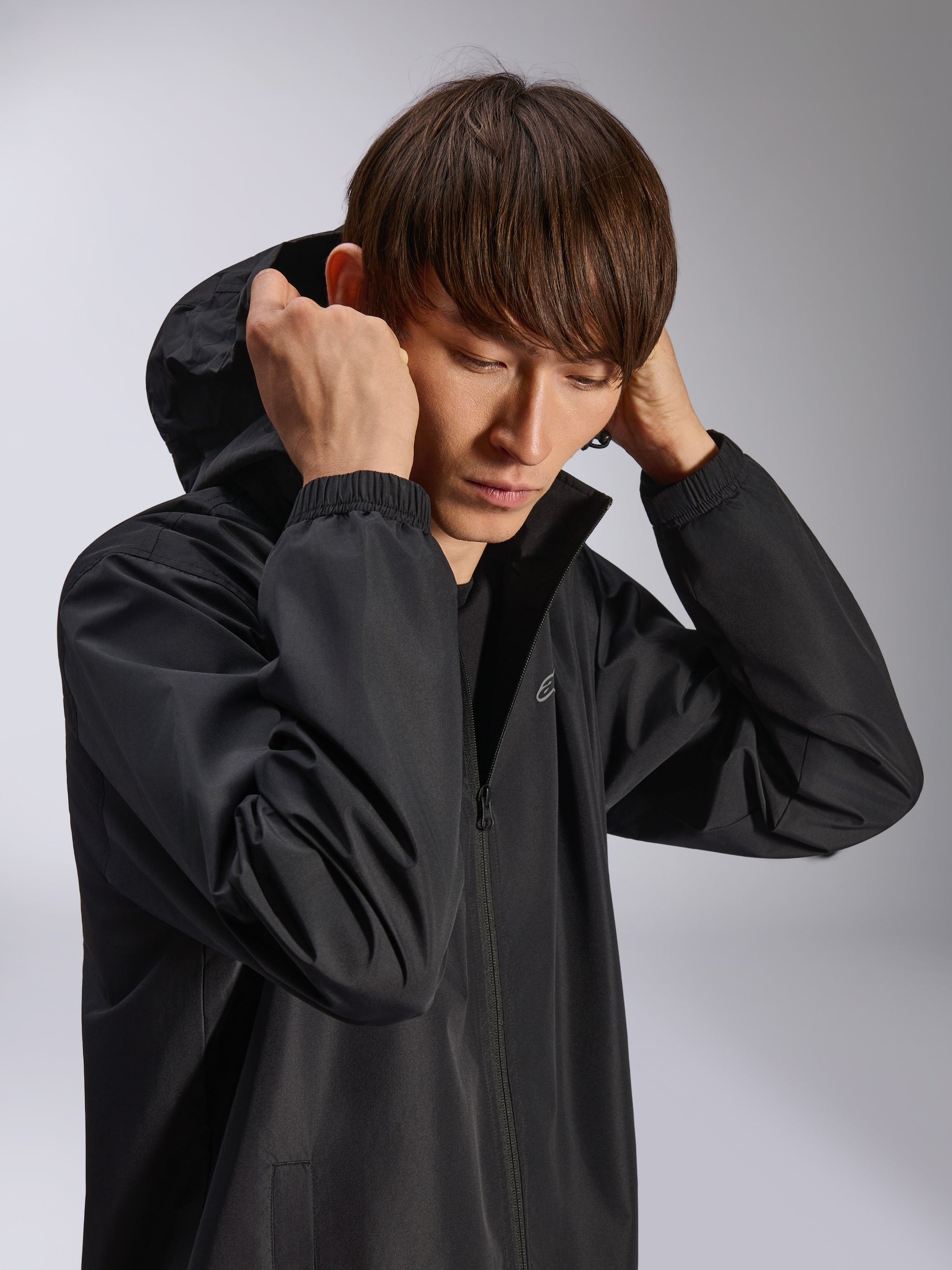 Alpinestars Overhaul Racer Jacket, Black hooded rain shell, worn by model with hood up, featuring water-resistant 2.5L fabric, elasticated cuffs, and minimal chest logo, casual motorsport-inspired outerwear
