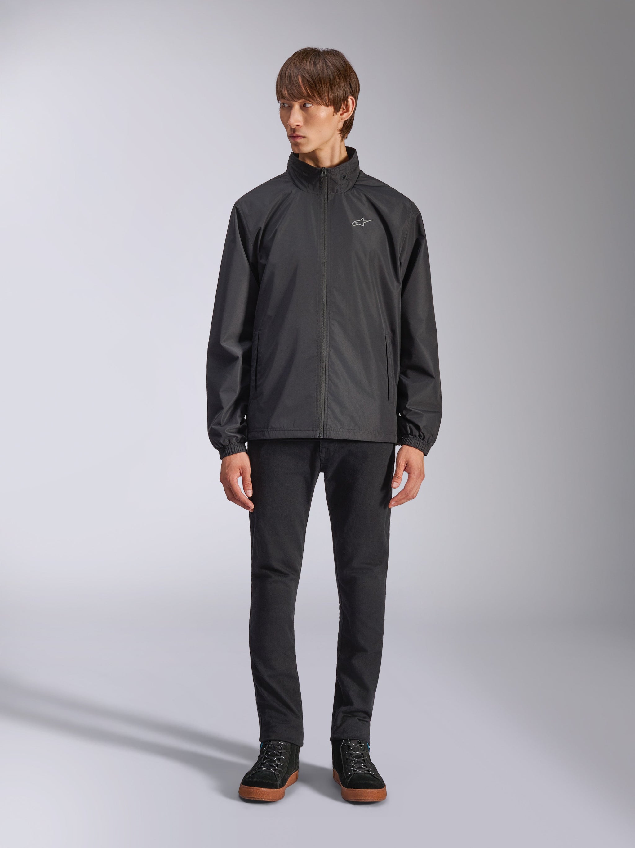 Alpinestars Overhaul Racer Jacket, Black Men's Rain Shell, full-length view on a model paired with black pants and casual sneakers, featuring a small white logo on the chest, water-resistant fabric, and casual motorsport-inspired design