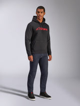 Alpinestars Blaze V3 Hoodie, black pullover fleece featuring a red Alpinestars logo on the chest, worn by a male model with dark denim jeans and black sneakers, casual motorsport-inspired streetwear