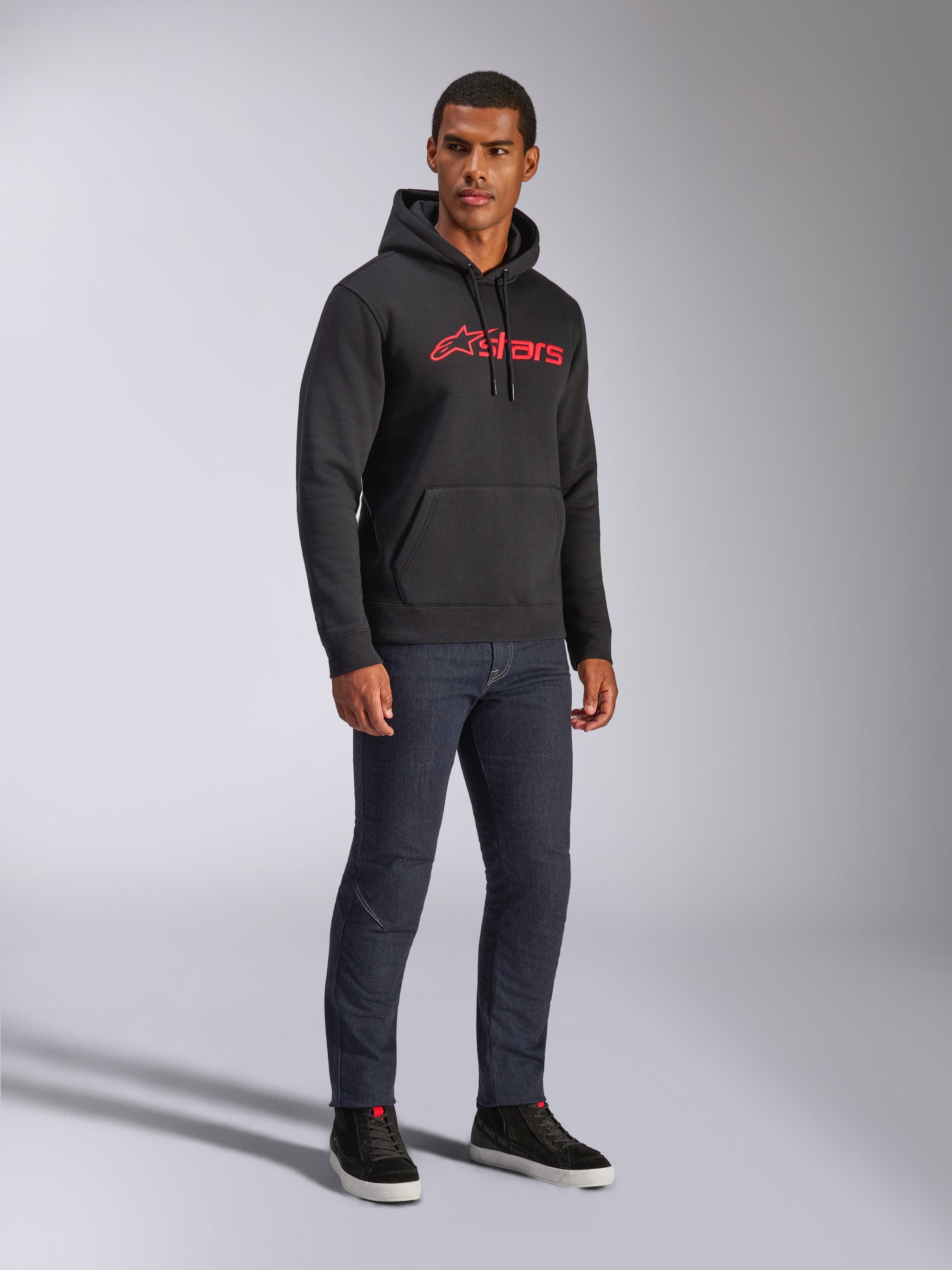 Alpinestars Blaze V3 Hoodie, black pullover fleece featuring a red Alpinestars logo on the chest, worn by a male model with dark denim jeans and black sneakers, casual motorsport-inspired streetwear