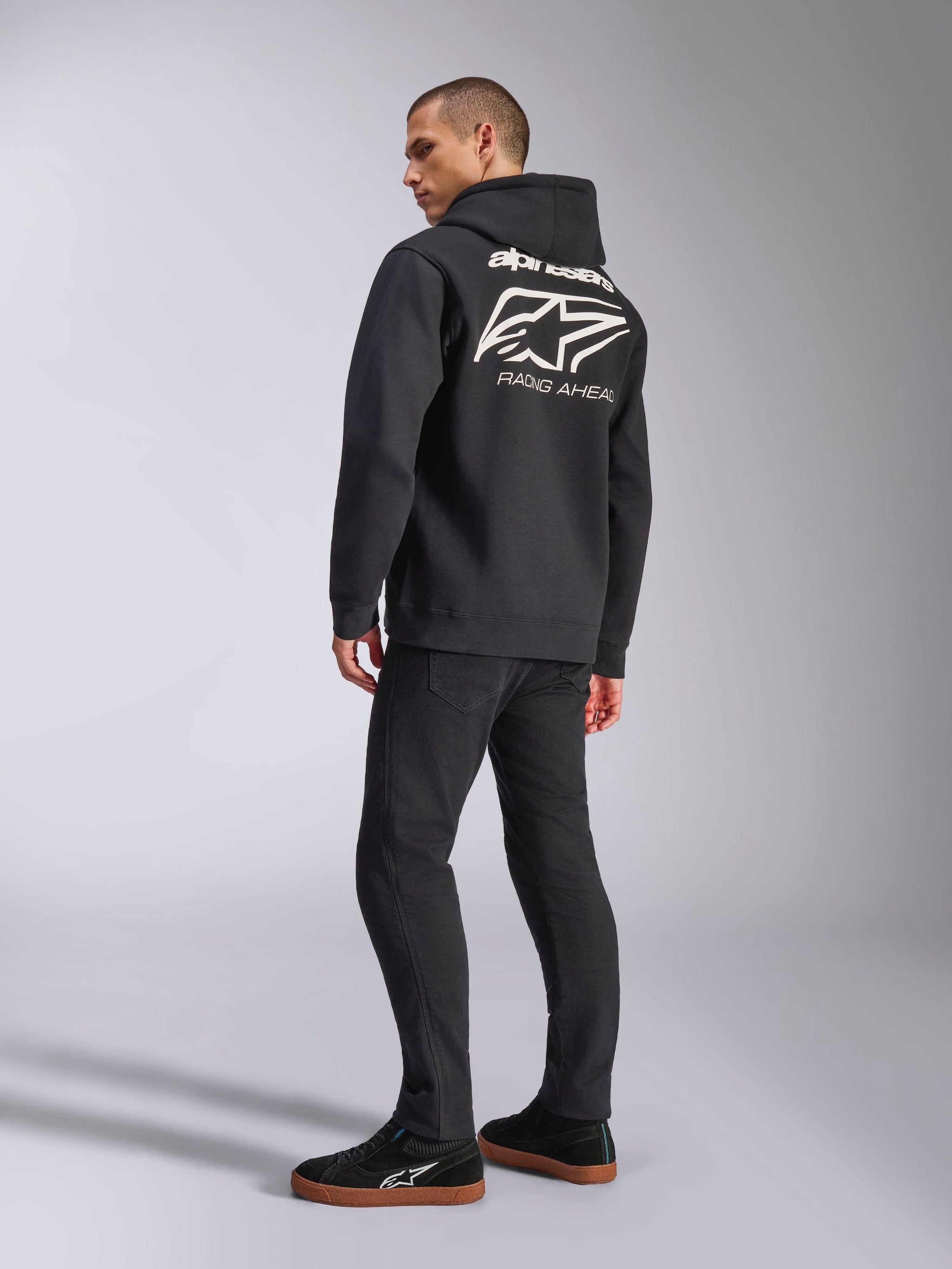 Model wearing Alpinestars Formulation Hoodie, Black, back view featuring large white Alpinestars logo and 'RACING AHEAD' wordmark, casual motorsport-inspired streetwear paired with black pants and black sneakers