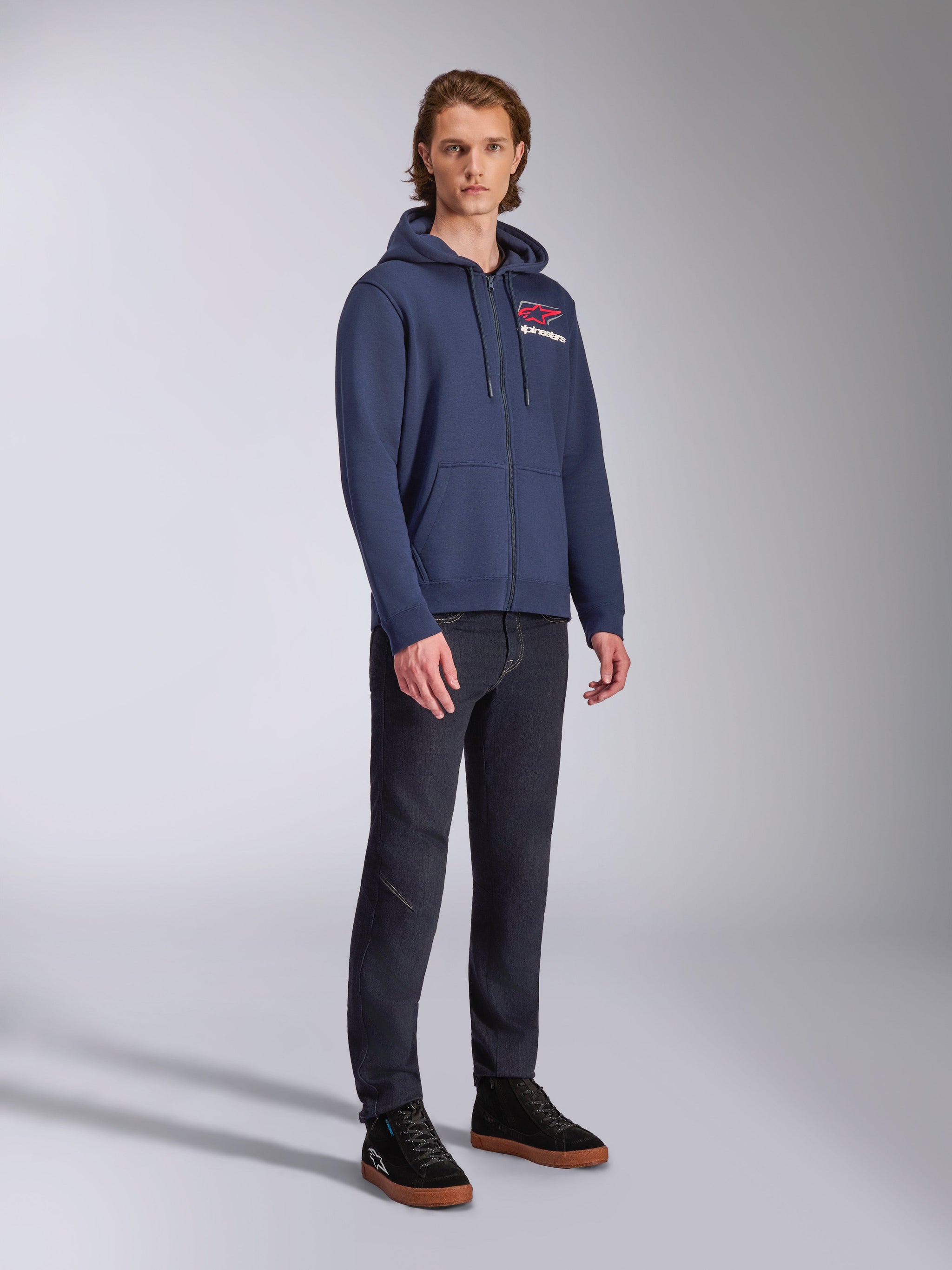 Male model wearing Alpinestars Formulation Hoodie, navy blue zip-front poly fleece, red and white logo on the chest, paired with dark jeans and black sneakers, casual motorsport-inspired streetwear