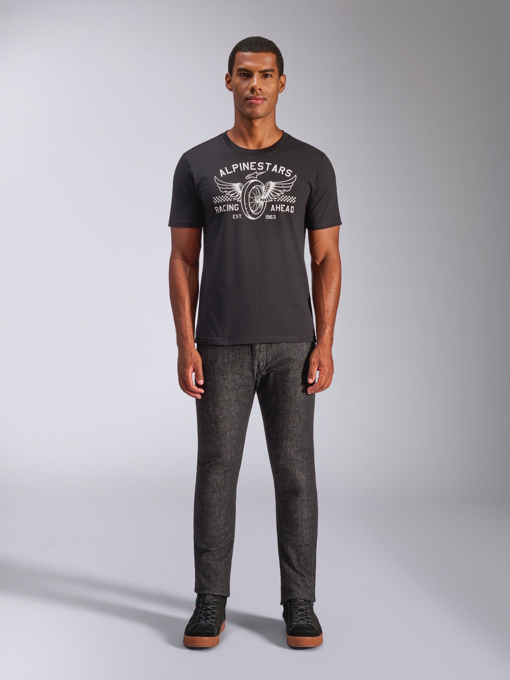 Alpinestars Heritage Patch CSF Tee, Short Sleeve T-Shirt, Black, featuring a white winged wheel graphic with 'Racing Ahead' text, worn by a model with dark grey pants and black sneakers, Casual Motorsport Streetwear