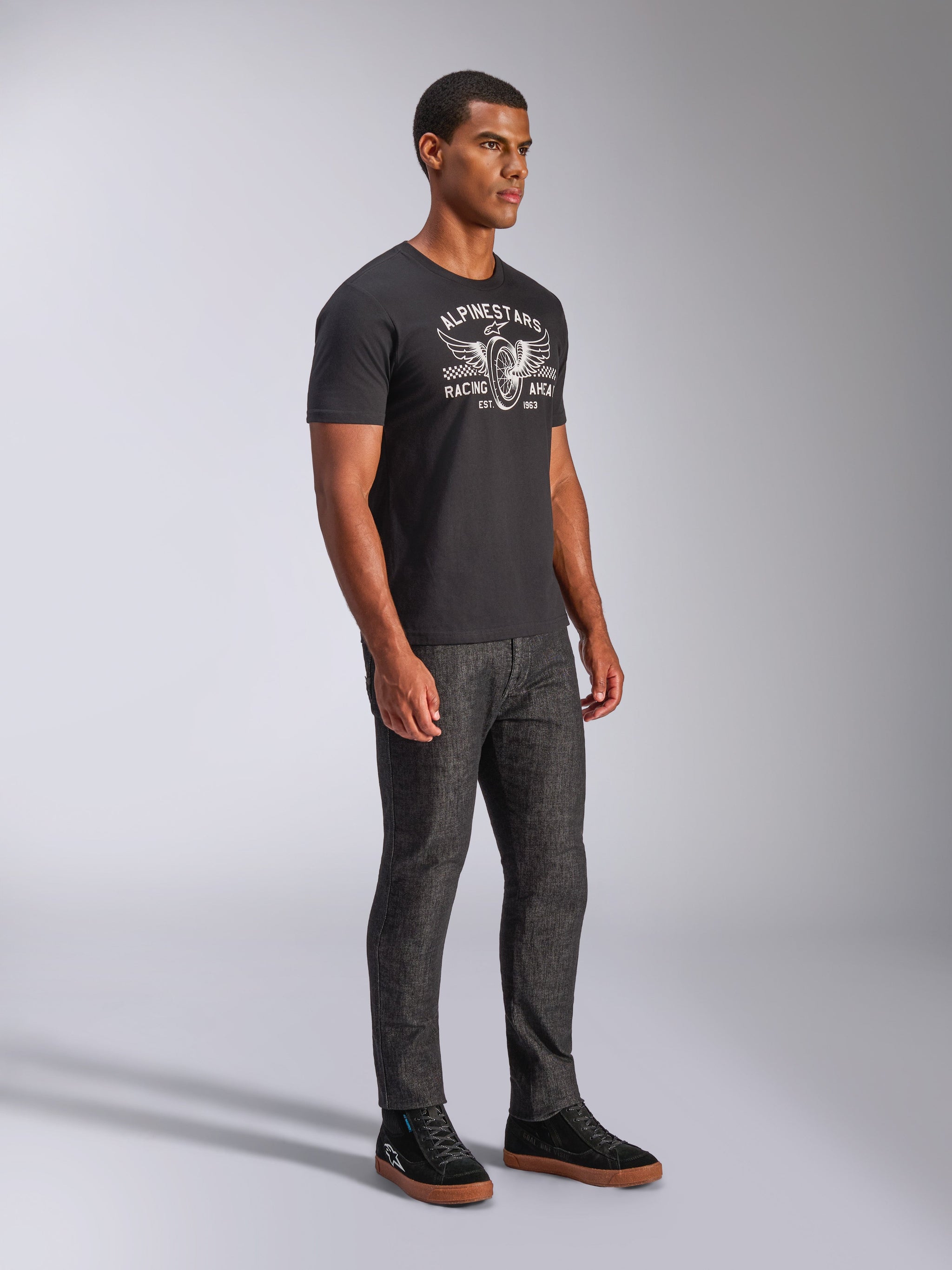 Alpinestars Heritage Patch CSF Tee, Short Sleeve T-Shirt, Black, White Winged Wheel 'Racing Ahead' graphic, worn by model with dark jeans and black sneakers, casual motorsport streetwear