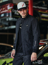 Alpinestars Precedent Jacket, black quilted jacket with Formula 1 styling, featuring an iconic angled quilt pattern, chest logo patch, and white sleeve embroidery, worn with a matching Alpinestars cap in a motorsport garage setting.