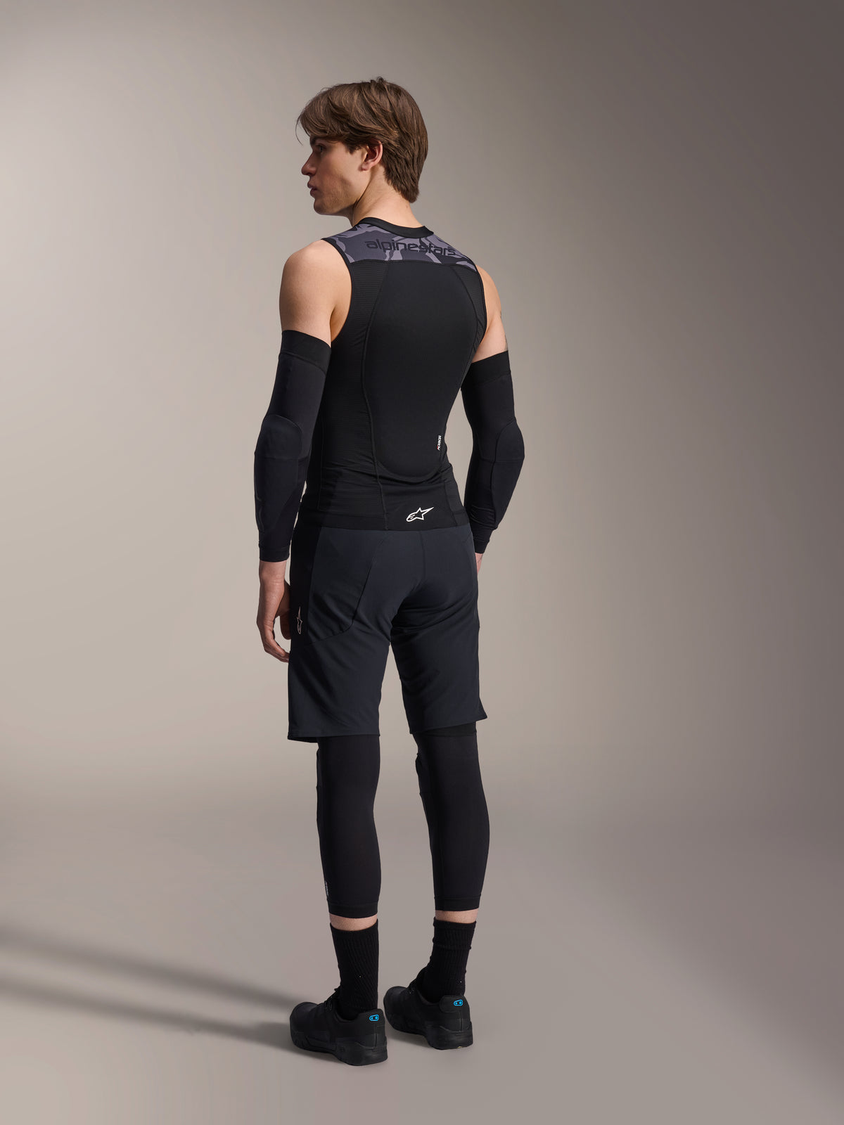 Male model from the back wearing Alpinestars A-Motion Plasma Protection Vest, Black Camo, sleeveless mesh design with integrated back protector, grey camo pattern on shoulders, worn with black elbow protectors and mountain bike shorts, professional cycling gear