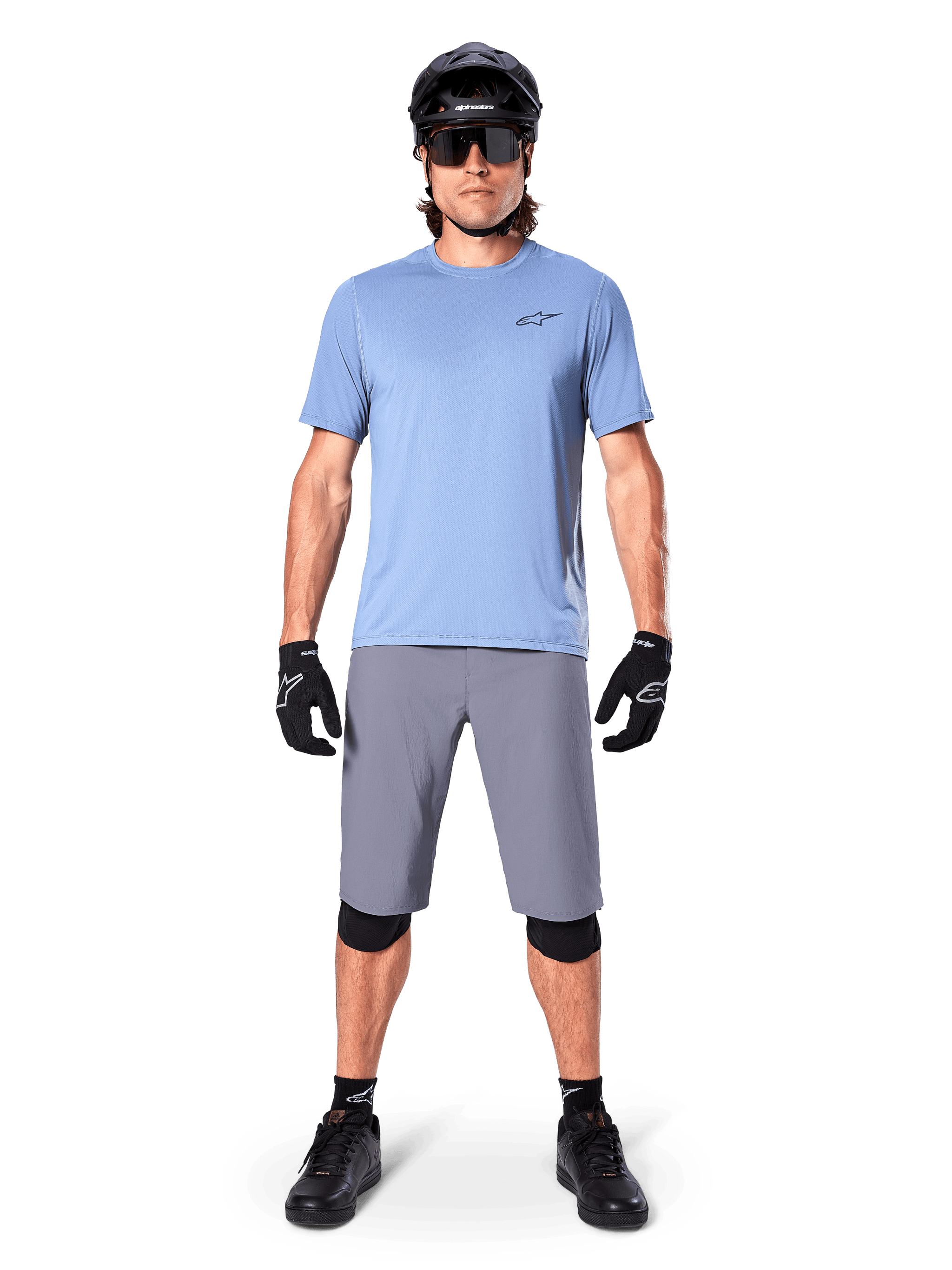 Male rider wearing Alpinestars A-Dura Shorts in Dark Gray, styled with a light blue short-sleeve t-shirt, black helmet, sunglasses, and black knee pads, complete mountain biking outfit for off-road performance.