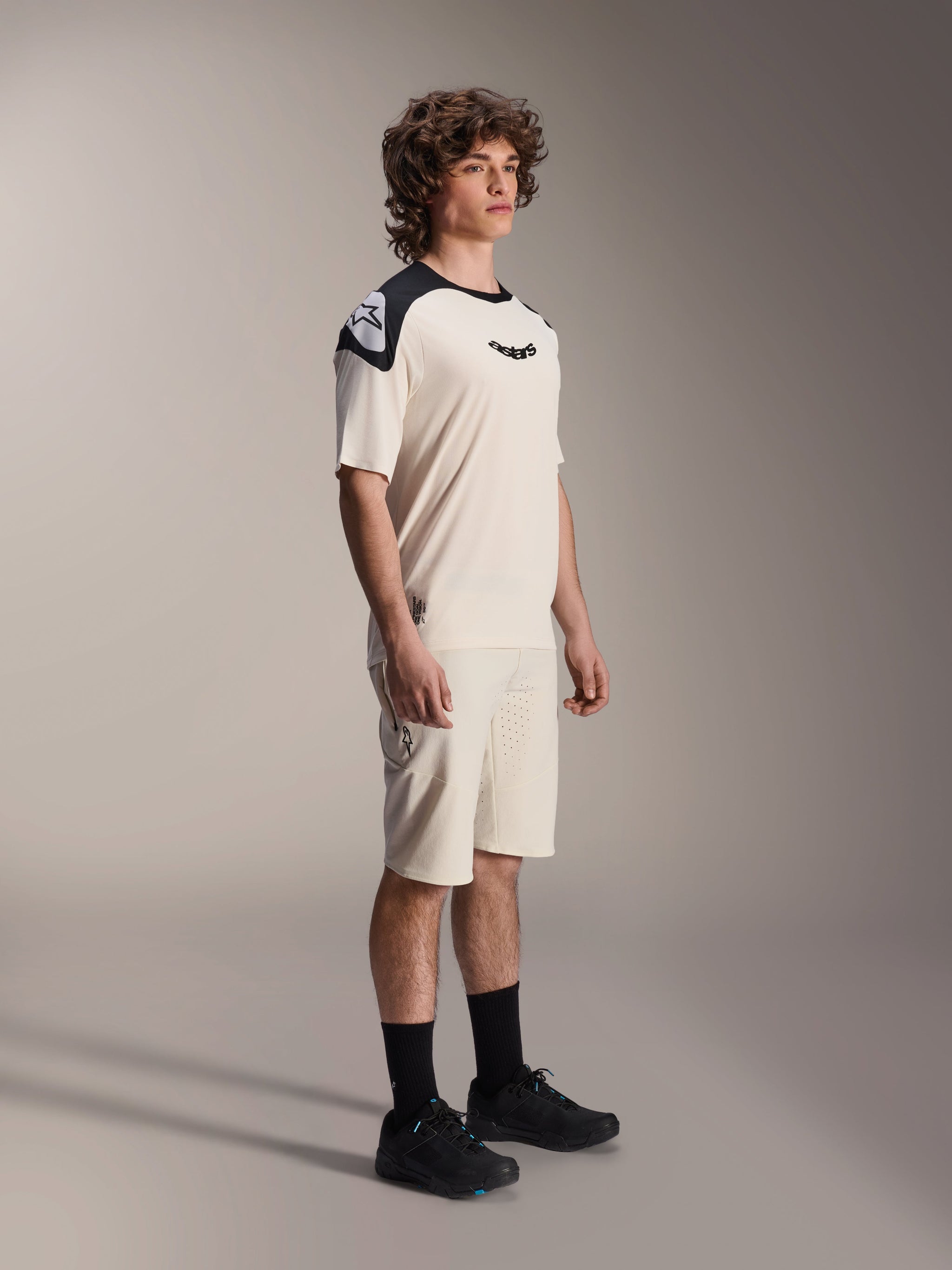 Male model wearing Alpinestars A-Aria Elite Shorts in Ecru, technical mountain bike shorts with laser-cut ventilation, paired with a matching cream and black short-sleeve jersey and black shoes, professional cycling apparel.