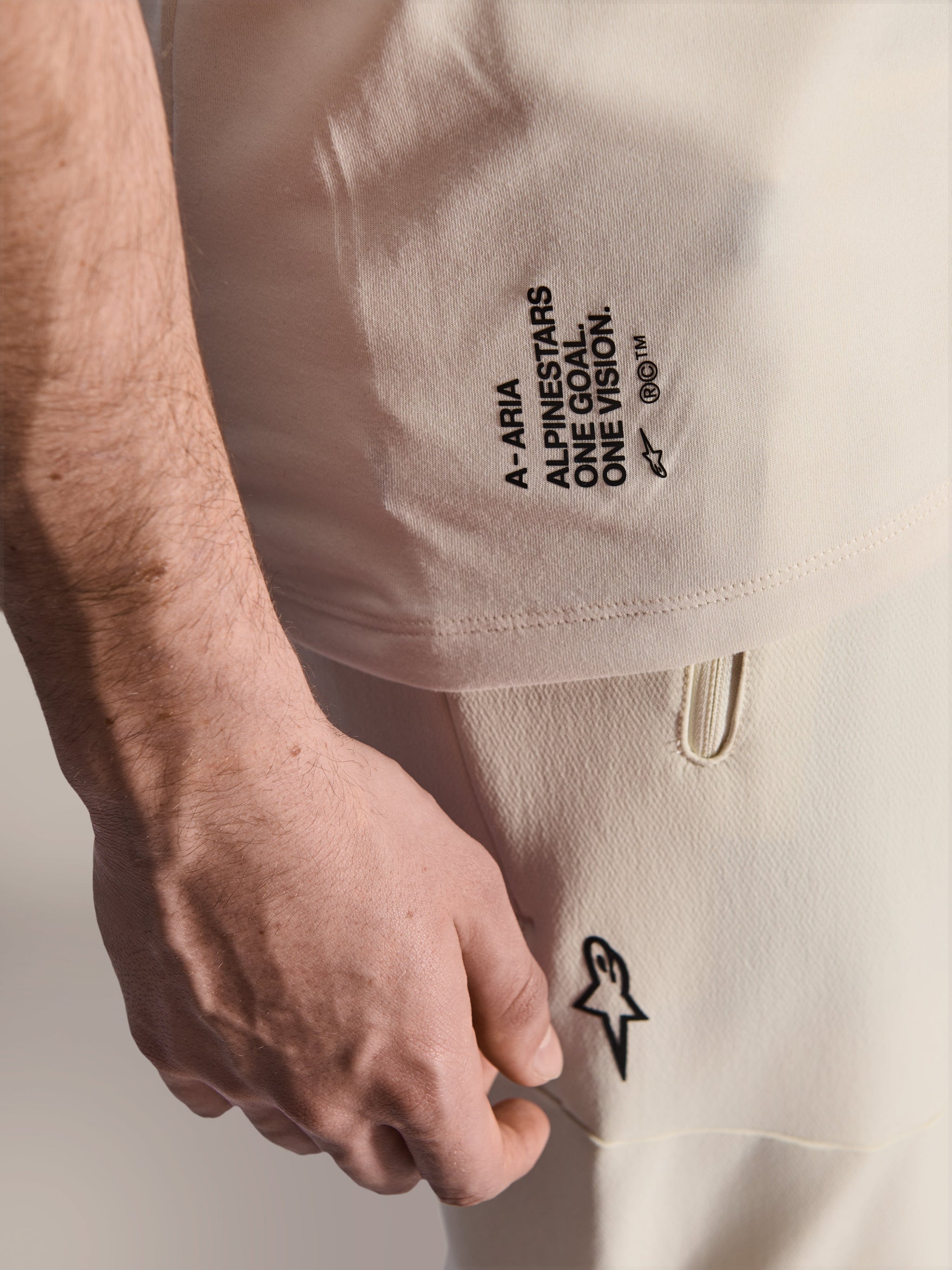 Alpinestars A-Aria Polartec Elite Short Sleeve Jersey, Ecru color, close-up of side branding featuring 'A-ARIA', 'ALPINESTARS', 'ONE GOAL. ONE VISION.', and Astars logo, worn with matching off-white pants, premium MTB performance apparel