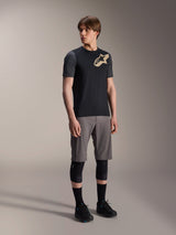 Male model wearing Alpinestars A-Dura Lipan Short Sleeve Jersey in Black, featuring dark grey sleeves and a tan Astars logo on the chest, paired with grey mountain bike shorts, black knee pads, and black cycling shoes, coordinated mountain bike riding gear