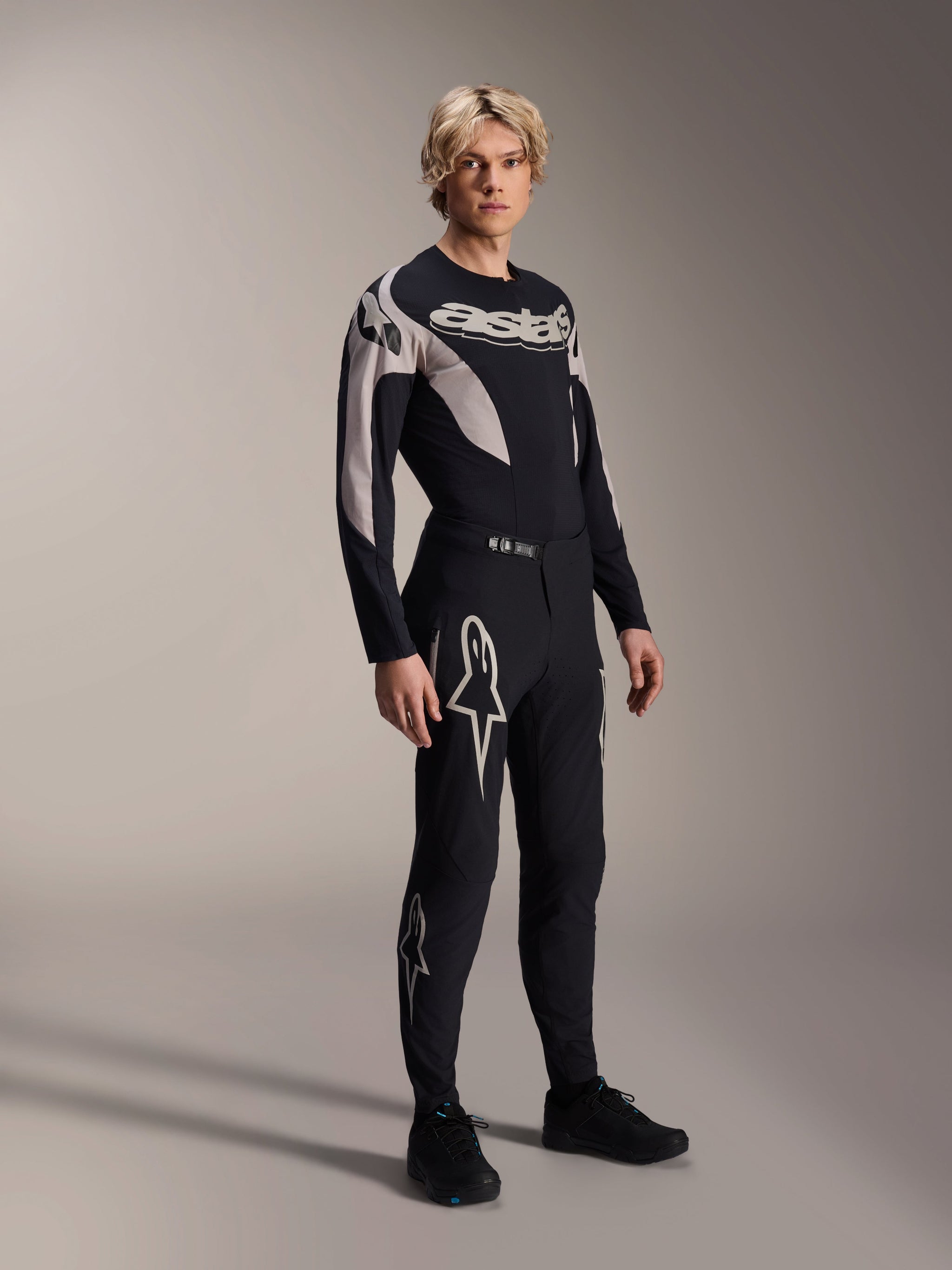 Male model wearing Alpinestars A-Supra Melt Jersey - Long Sleeve, Black with grey graphic accents, 'astars' chest logo, paired with matching black pants and shoes, performance mountain bike kit for off-road racing