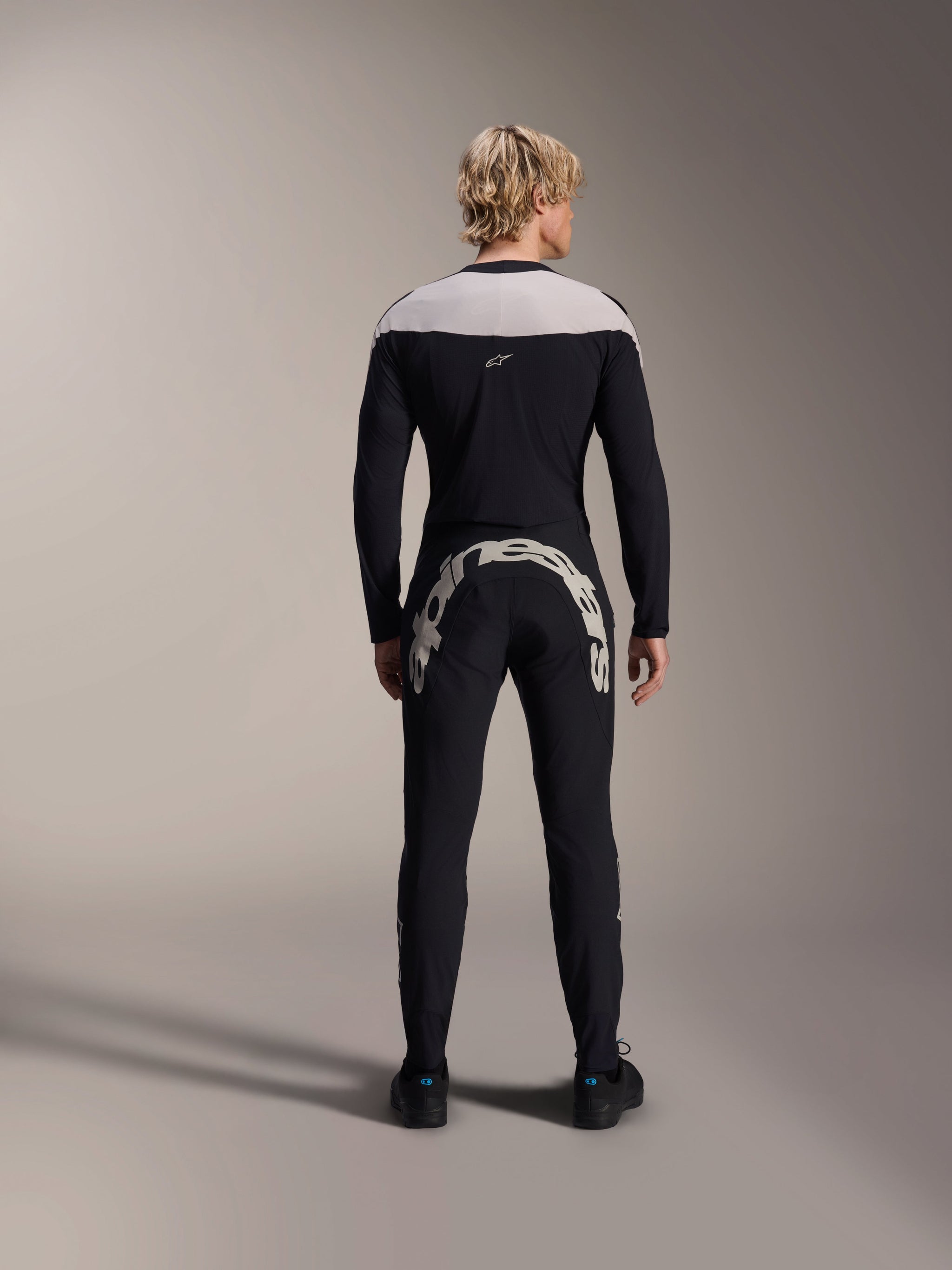 Man seen from behind wearing Alpinestars A-Supra Melt Long Sleeve Jersey in Black, light grey shoulder panel, paired with black MTB pants featuring a large Alpinestars logo across the rear, performance mountain bike kit