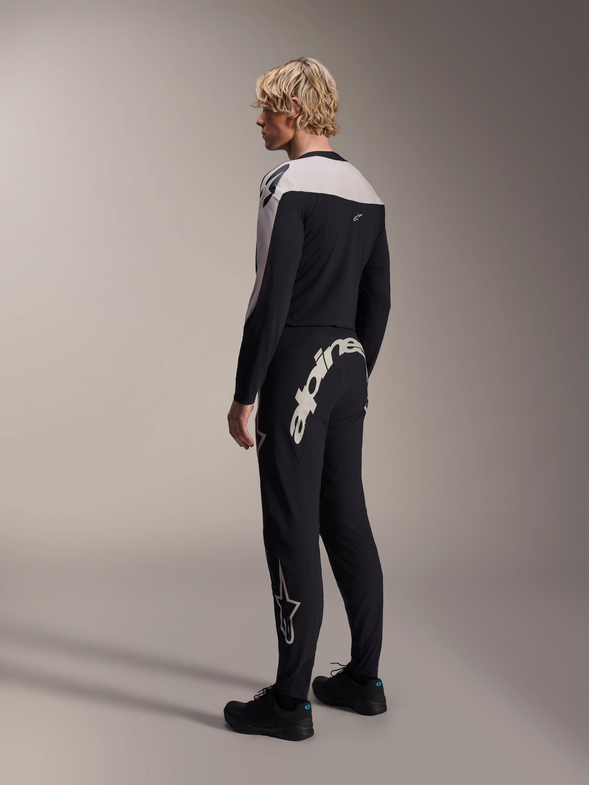 Alpinestars A-Supra Melt Long Sleeve Jersey, Black with white shoulder panels, worn by a model from the back and side, paired with black mountain bike pants featuring a large white wordmark, performance MTB racing kit