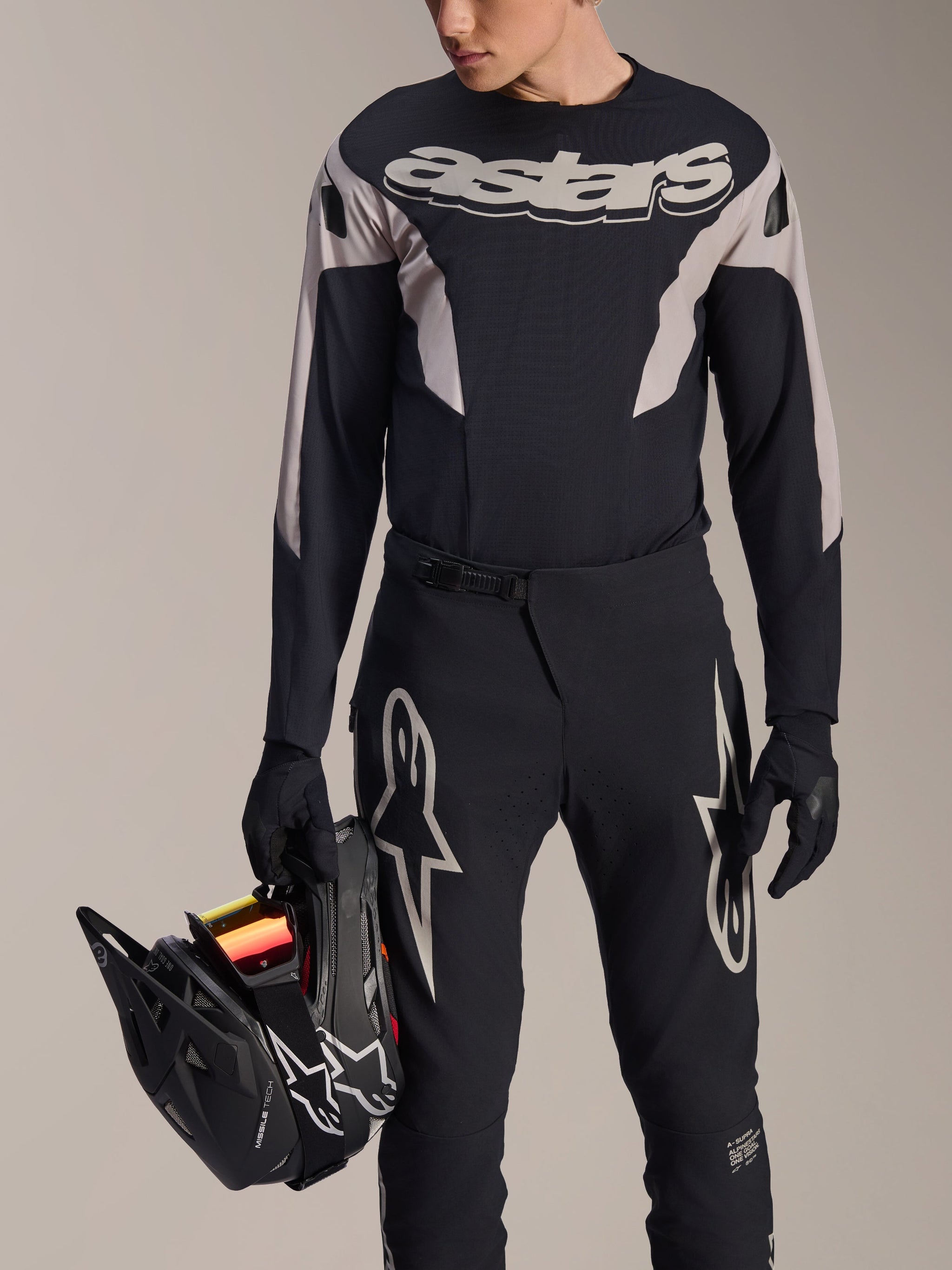 Alpinestars A-Supra Melt Long Sleeve Jersey, Black with grey graphic panels and bold 'astars' wordmark on chest, worn with black mountain bike pants and gloves while holding a black full-face helmet, performance MTB racing kit