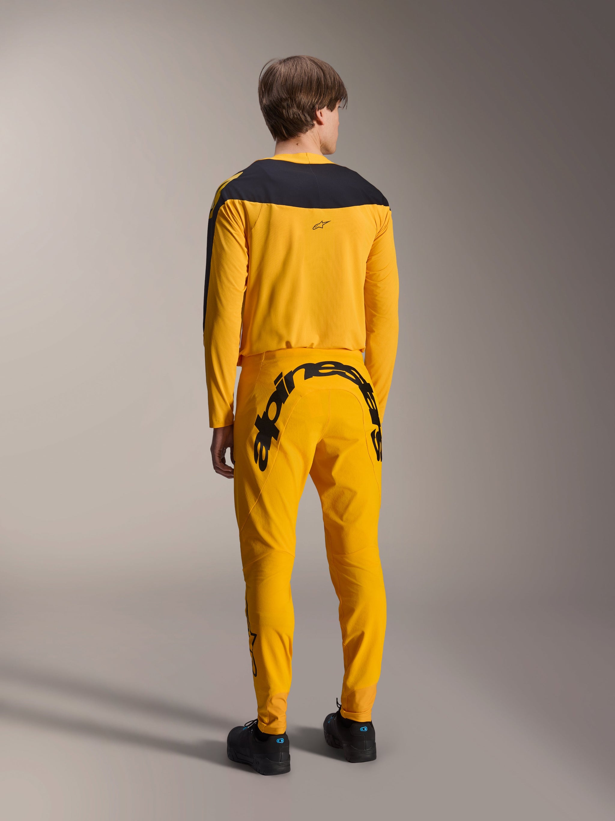 Alpinestars Limited Edition A-Supra Melt Gold Jersey, Long Sleeve MTB Jersey, Dark Yellow with black shoulder panel, worn with matching yellow pants featuring bold black branding on the back, rear view of performance cycling kit