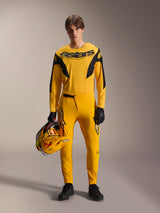 Male model wearing the Limited Edition A-Supra Melt Gold Gear-Set, Dark yellow long-sleeve jersey with black Astars logo, matching yellow pants with black accents, holding a yellow and black helmet, black gloves and technical shoes, mountain bike racing outfit
