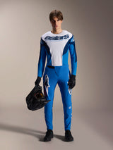 Male rider wearing Alpinestars A-Supra Melt Jersey - Long Sleeve in Blue and White, featuring 'astars' branding on the chest, matching blue pants, black gloves, and holding a black helmet, performance-focused MTB racing kit.