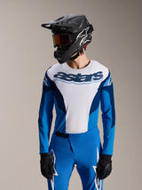Model wearing Alpinestars A-Supra Melt Long Sleeve Jersey in Blue and White, featuring 'astars' wordmark on the chest, styled with matching blue MTB pants, black full-face helmet, and goggles, complete mountain bike racing outfit.