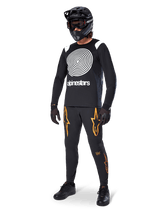 MTB rider wearing Alpinestars A-Supra Oscar Long Sleeve Jersey in Black, featuring the white vintage Oscar spiral graphic, black pants with orange star logos, black full-face helmet with goggles, and black gloves, professional mountain biking gear