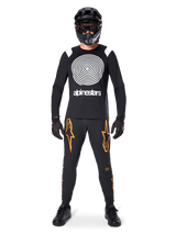 Male model wearing complete mountain bike gear, Alpinestars A-Supra Oscar Long Sleeve Jersey in black with white spiral chest graphic, black pants with orange star logos, black full-face helmet and goggles, black gloves and shoes, coordinated outfit for MTB gravity riding