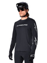 Male cyclist wearing Alpinestars A-Dura Switch Long Sleeve Jersey, Black with white wordmark and star logos on sleeves, styled with black MTB helmet and sunglasses, high-performance cycling gear for off-road riding