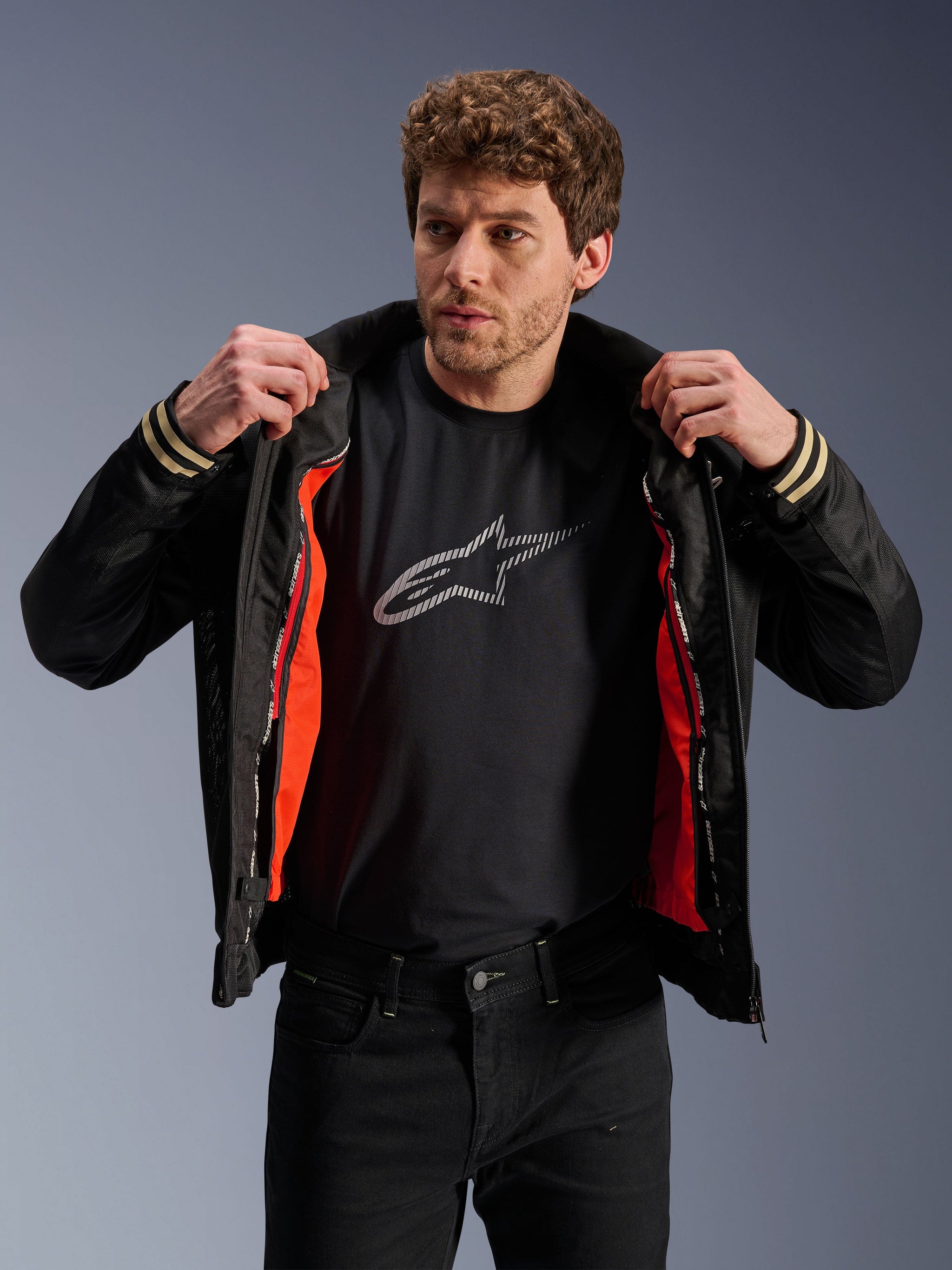 Man wearing Alpinestars Ignite Air Jacket, Black, featuring a mesh body and bright orange detachable windproof liner, paired with a black Astars logo t-shirt and black pants, urban contemporary motorcycle style