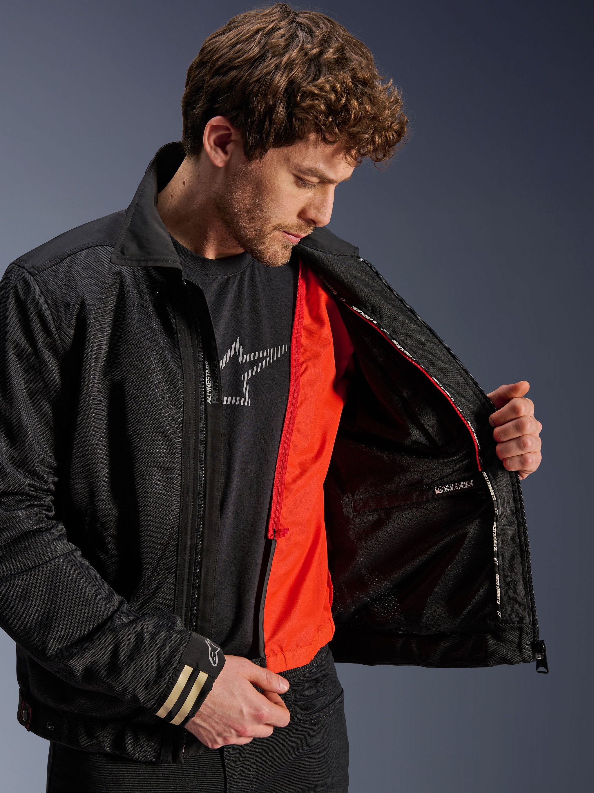 Alpinestars Ignite Air Jacket, Black Urban Motorcycle Jacket, featuring a bright orange detachable windproof liner and mesh body, interior view showing waterproof compartment, worn with a black Alpinestars logo t-shirt.