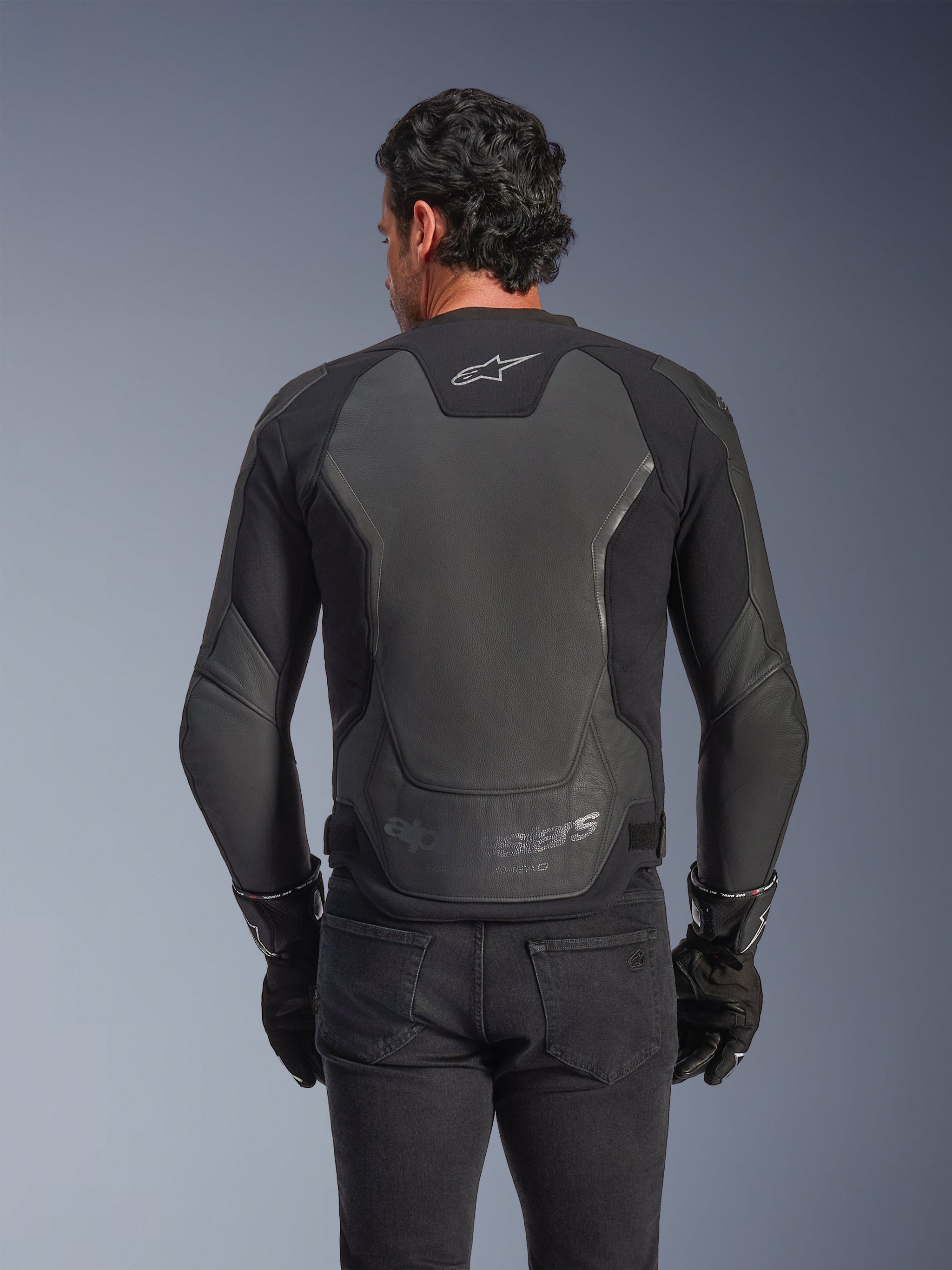 Alpinestars GP Force V2 Leather Jacket, Black, Rear view of a man wearing the jacket with black gloves and jeans, featuring large leather protective panels on the back, Alpinestars logo on the upper back, and embossed branding at the lower waist, sport-inspired motorcycle riding gear